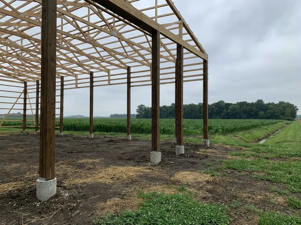 A post-frame home under construction, showing laminated timber columns on concrete piers and wide-span roof trusses with no interior bearing walls.