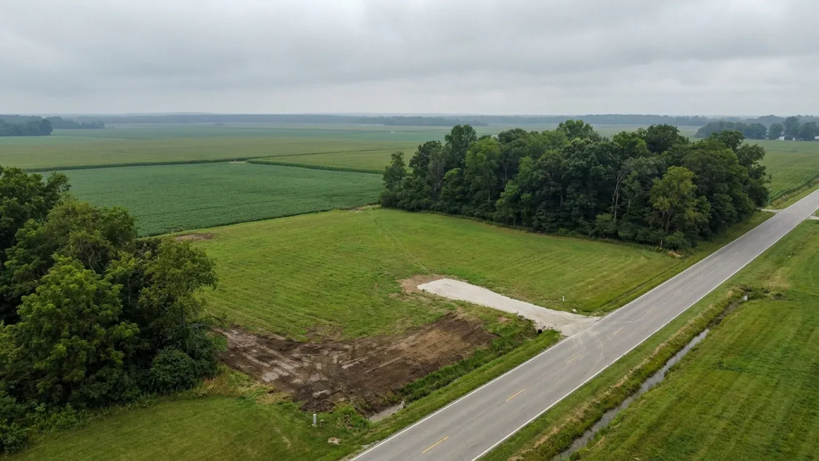 Aerial view of an undeveloped rural lot beside crop fields, with mown grass, a hardwood woodlot along one side and a short gravel drive in from the road.